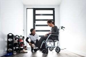 "Caregiver assisting an elderly woman in a wheelchair near the entrance of a modern home. The caregiver helps with the woman's shoes, emphasizing transportation issues and the challenges wheelchair users face in daily life. The scene highlights the importance of accessible home environments and supportive care for individuals with mobility impairments.