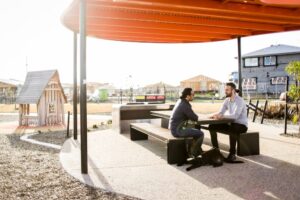 Two men sitting at an outdoor picnic table under a modern orange shade structure, discussing transportation issues. The setting includes a small wooden playhouse and residential construction in the background, highlighting a community park. A black guide dog rests at the feet of one of the men, emphasizing accessibility and transport challenges for wheelchair users.