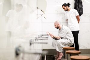 Two men engaged in a fatigue management plan: one seated and washing his hands, reflecting a calming routine, while the other stands nearby, offering support. The setting is a clean, tiled room with stools and faucets, highlighting a structured environment aimed at managing fatigue effectively.