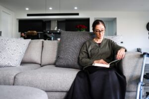 Elderly woman reading a book on a grey sofa in a modern living room, with a walker positioned nearby. The scene illustrates transportation issues for wheelchair users, emphasizing the need for accessible home environments. The background features a sleek kitchen area with people engaged in conversation, highlighting the everyday challenges faced by individuals with mobility impairments.