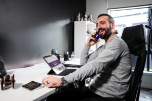 Man discussing fatigue management plans over the phone while seated at a modern home office desk, equipped with a tablet, speakers, and a motivational book. The professional setting, including a large window and personal mementos, emphasizes the importance of effective fatigue management strategies in maintaining productivity.