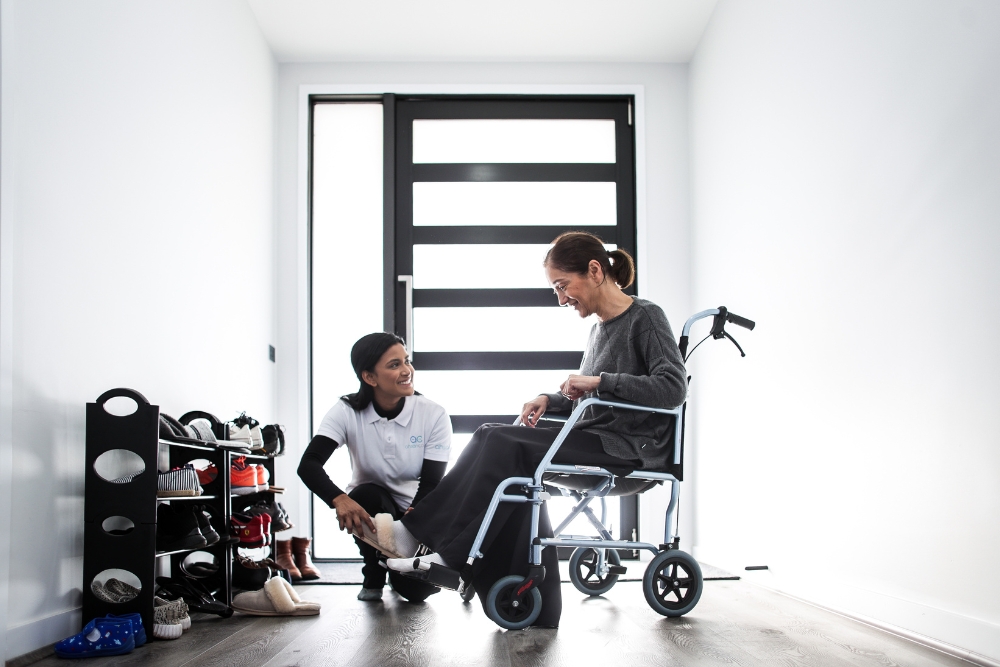 A caregiver assists a woman in a wheelchair with putting on shoes in a brightly lit hallway. The caregiver is kneeling beside the woman, who is smiling, as she receives help with personal care tasks. A shoe rack filled with various shoes is visible in the background. The image illustrates the importance of support with personal care tasks for individuals with mobility challenges.