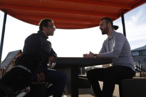 Two men discussing NDIS for mental health outdoors, sitting at a table under a canopy, with a guide dog in the foreground.