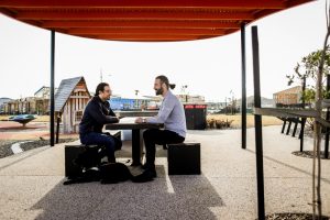 Two men sit across from each other at an outdoor table, engaged in a discussion about technology for disabled people.