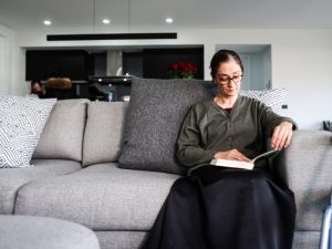 A woman reading a book on a grey sofa in a modern living room, representing NDIS transport assistance at home.