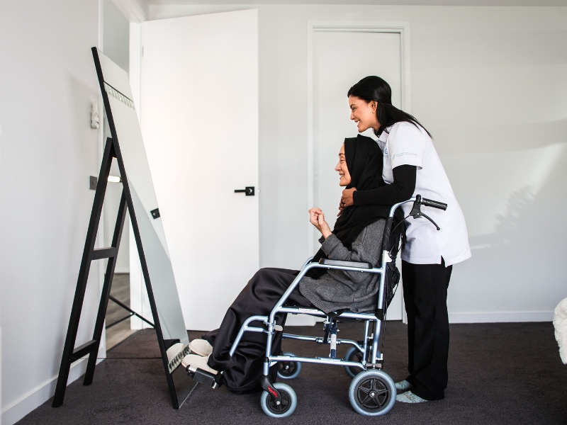 Healthcare worker providing assistance for disabled persons, helping a woman in a wheelchair in front of a mirror.