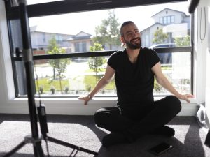 Man seated cross-legged by a window, embodying relaxation and innovative community participation in casual, comfortable settings.