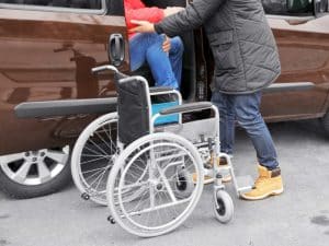 A travelling companion helping a client in a wheelchair exit a car with care and support.