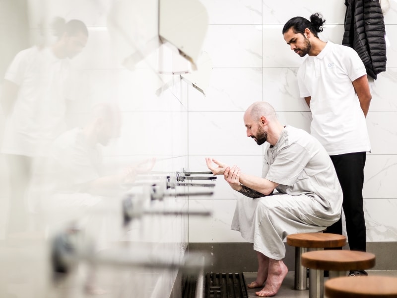 A support worker assists a man in a communal washroom, demonstrating pain assessment and disability care in daily routines.