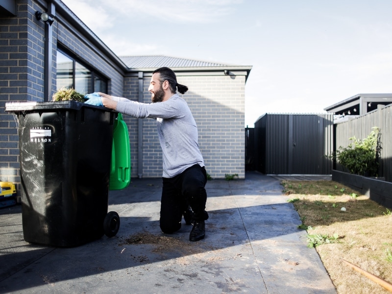 A man wearing gloves kneels beside a 240L bin, safely disposing of garden waste, ensuring NDIS rubbish removal safety.