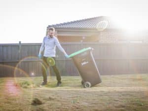 A man wearing gloves safely moves a 240L green-lid bin in a backyard, demonstrating NDIS rubbish removal safety.