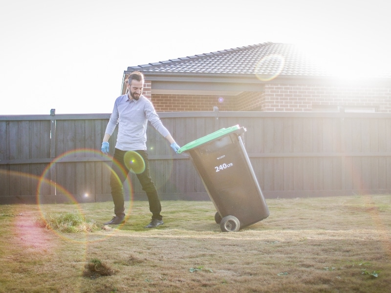 A person wearing a light blue long-sleeved shirt, dark pants, and light blue gloves is pulling a large, dark green and black wheeled bin with a light green lid across a grassy lawn. The bin has "240Ltr" printed on its side, suggesting its capacity. This depicts contaminated waste removal from a residential area, indicated by the house and fence in the background. The scene is brightly lit with sunlight causing lens flares.