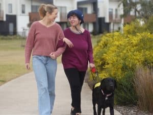 Two women walking with a guide dog during ndis community events in a suburban neighbourhood.