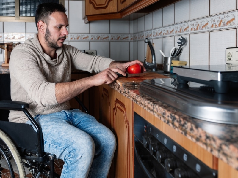 Man in wheelchair slicing tomato at bench as part of household tasks assistance in the kitchen.